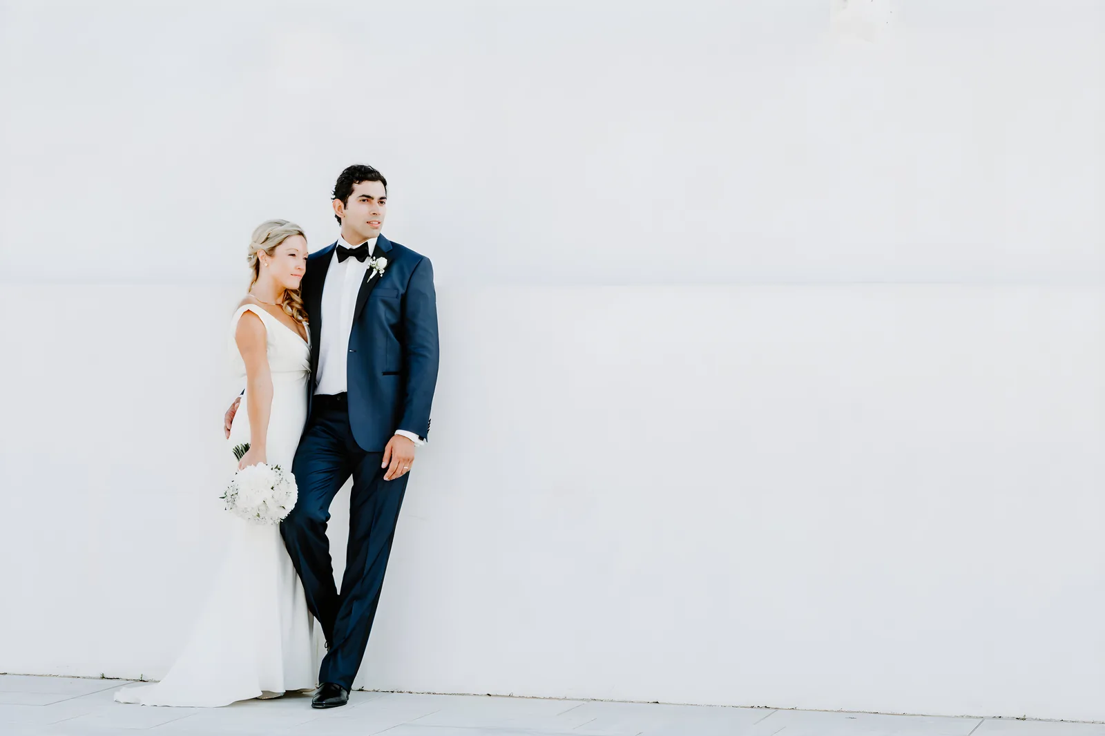 Bride in a white gown and groom in a navy tuxedo standing against a pale wall