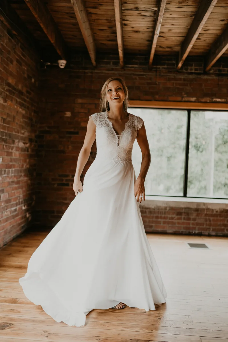 A bride laughing as she lifts her gown in a brick-walled room