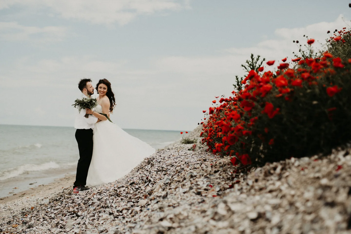Bride and groom on a pebble beach beside a bank of red poppies