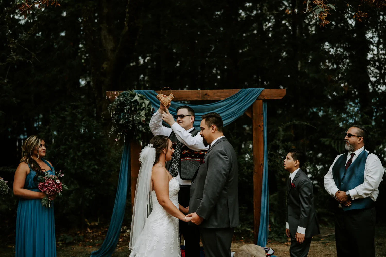 A ceremony beneath a wooden arch draped in blue fabric, while a boy in a suit watches from the side