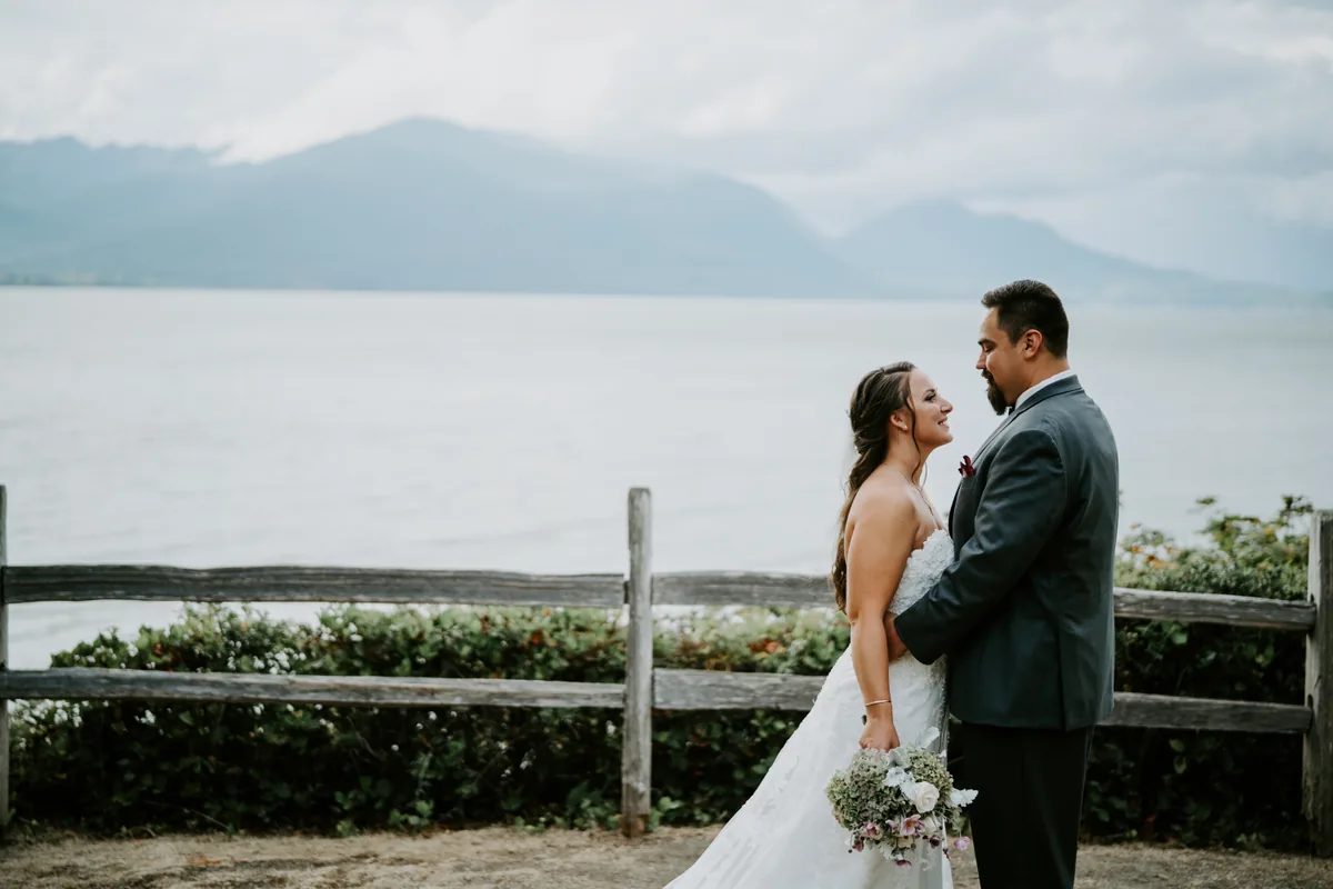 Bride and groom by a wooden fence, with a lake and mountains behind them