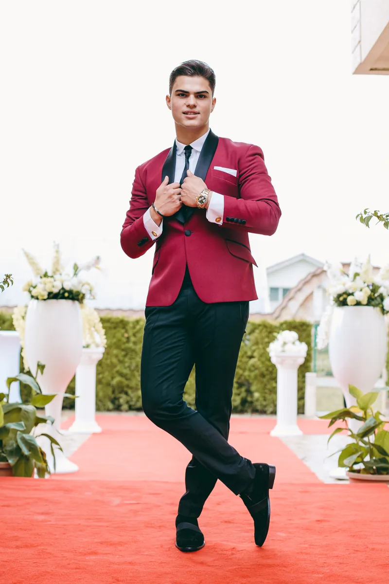 A groom in a burgundy jacket standing on a red carpet