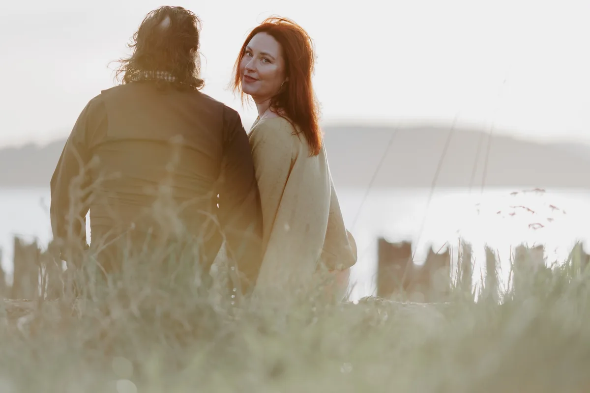 A woman looks back over her shoulder while sitting close to her partner in tall grass by the water